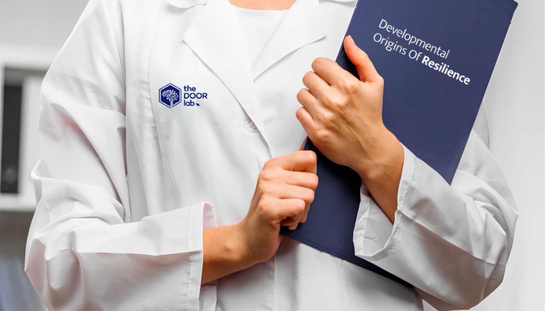 The DOOR Lab logo embroidered on a researcher's lab coat alongside a Developmental Origins of Resilience book.